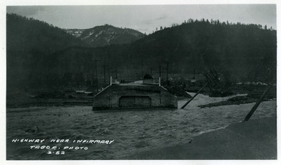 Floodwaters approach highway near infirmary. A bridge appears to have been blocked by the floodwaters and several telephone poles appear damaged. Two people are standing on the bridge.
