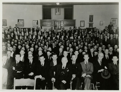 Men and marching band members gather for a photograph in celebration of Aerie #54 Fraternal Order of Eagles' 34th anniversary meeting. A welcome sign hangs between two American flags in the background.