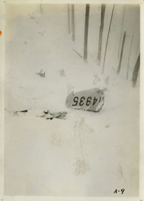 Debris from an airliner crash partially covered in snow. Numbers "4935" can be seen on the remnant. A few bare trees in the background.