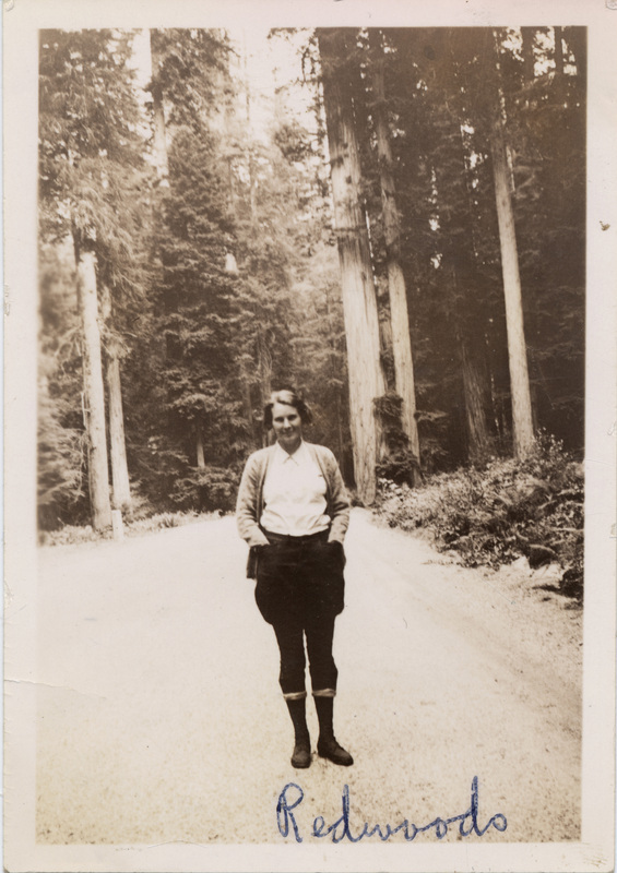 A woman stands on a road in front of trees and smiles at the camera. "Redwoods" is written on the photograph. The number 51 is stamped on the back of the photograph.