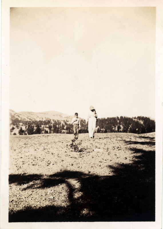 Two people walk over a rocky area, with pine trees in the background. The number 46 is stamped on the back of the photograph.