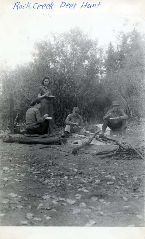 Four people sit around the ashes of a campfire and eat from plates. The photo is captioned: "Rock Creek Deer Hunt." The number 213 is stamped on the back of the photograph.