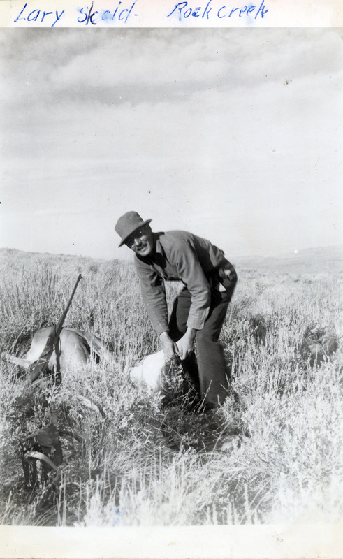 A man stands over a dead deer with a rifle leaning on it. He is lifting up part of the deer and smiling at the camera. The photo is captioned: "Larry Skold - Rock Creek." The number 213 is stamped on the back of the photograph.
