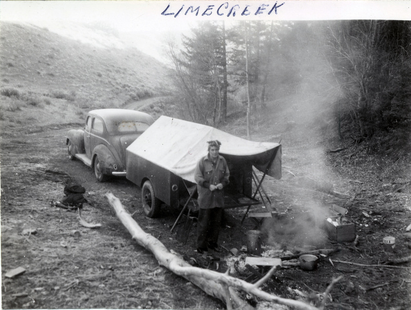 A car towing a camping trailer is parked in the woods. Next to it, a person stands in front of a campfire. The back of the photograph is stamped with: 874 [illegible].
