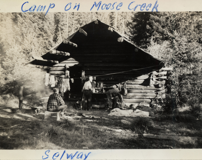 Three people sit and stand around a log shelter. There is smoke from a fire and clothes are hanging from a line. The photo is captioned: "Camp on Moose Creek Selway." The back of the photograph is stamped with the number 199 and "Printed by The Camera Shop Sun Valley, Idaho Oct 15 1942."