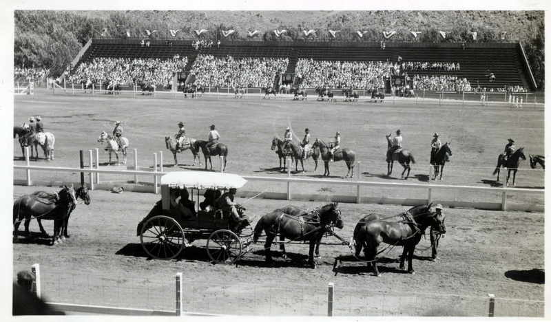 A team of horses pull a carriage in front of a rodeo arena.