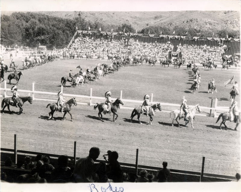 A group of people wearing feathered head coverings ride horses in front of the rodeo arena. The picture is captioned "rodeo."