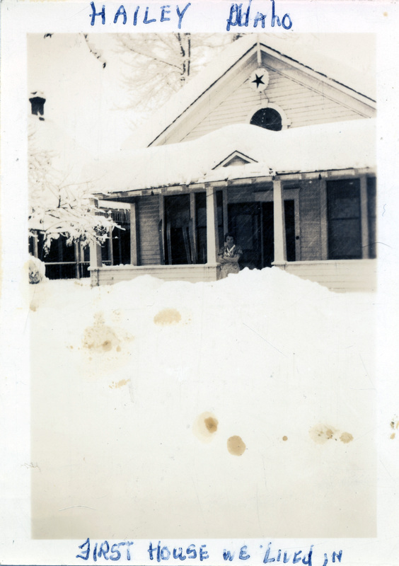 A woman stands on the porch of a one-story house covered in snow. "Hailey Idaho, first house we lived in" is written on the front of the photo. The back of the photo is stamped with the number 716.