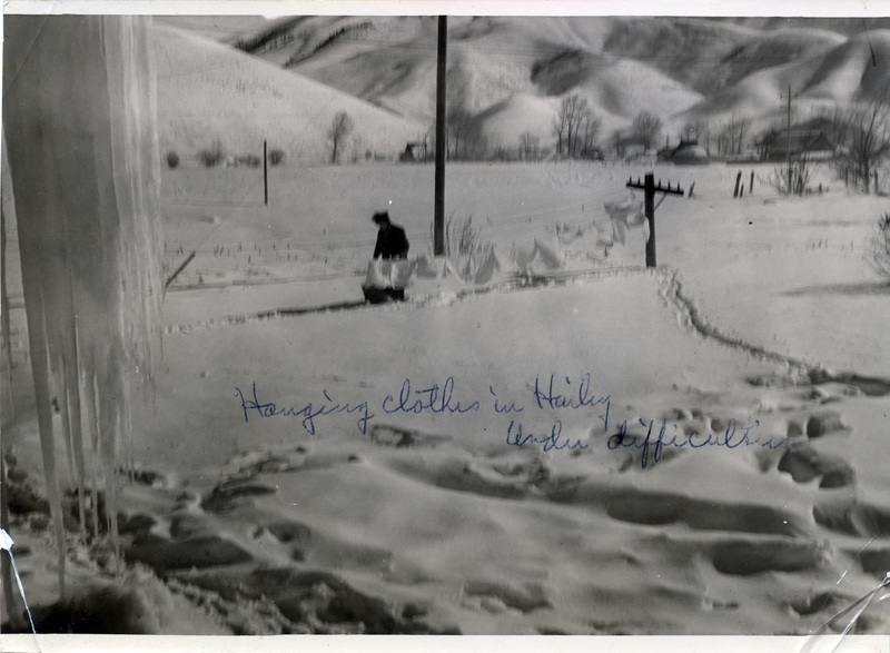 A person hangs clothing on a clothesline that stands in a snowy field. "Hanging clothes in Hailey under difficulties" is written on the front of the photo.