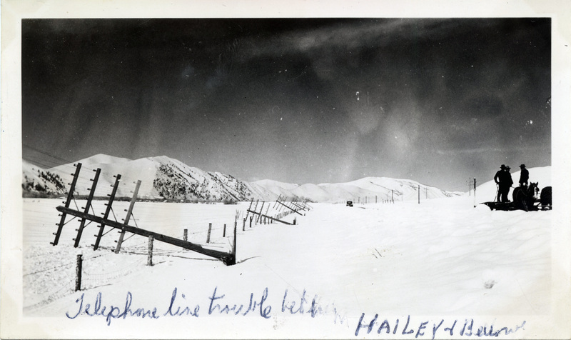 A group of people stand near a telephone pole, which has fallen down in the snow. "Telephone line trouble better in Hailey & Bellevue" is written on the front of the photograph. The back of the photograph is stamped with the number 16.