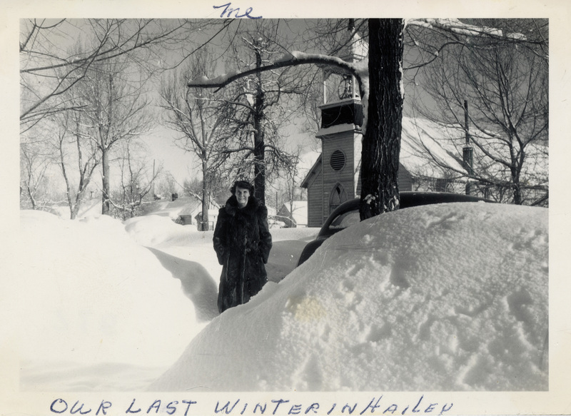 A woman stands in the snow, in front of a building with a bell tower. "Me, our last winter in Hailey" is written on the front of the photo. The back of the photo is stamped with the number 716.