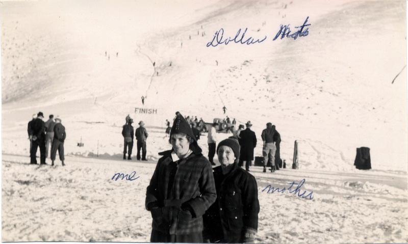 Two women pose together in front of a ski hill. People in the background ski towards a Finish sign at the bottom of the slope. "Dollar Mt., me, mother" is written on the photograph.