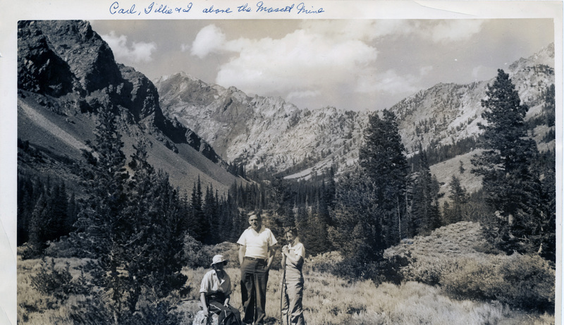 Three people pose together in a forest. A mountain range is visible in the background. "Carl, Tillie, and I above the Mascot Mine" is written on the front of the photograph.