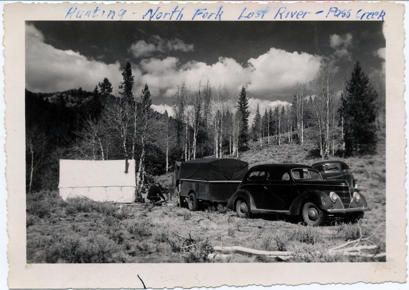 Two automobiles are parked next to a small structure in a forested area. A man tends to some equipment next to a trailer pulled by one of the automobiles. "Hunting - North Fork Lost River - Pass Creek" is written on the front of the photograph.