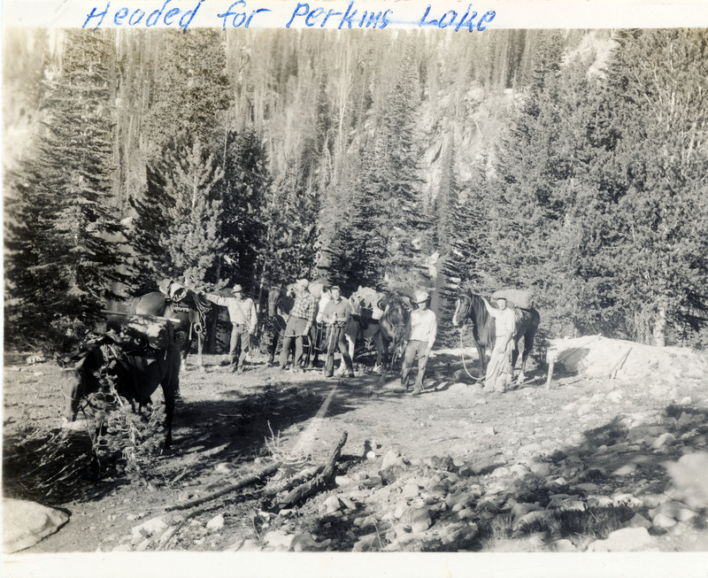 A group of people pose with horses in a forest area. "Headed for Perkins Lake" is written on the front of the photograph.