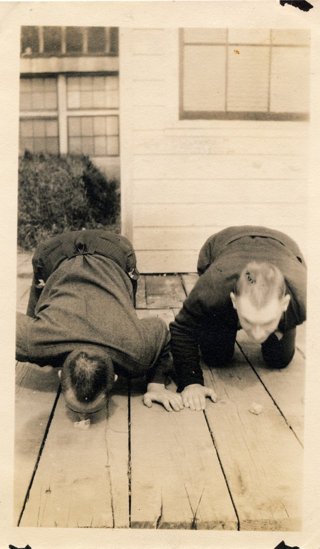 Two men are next to each other, on all fours, on a deck. The back of the photograph is stamped with the number 16