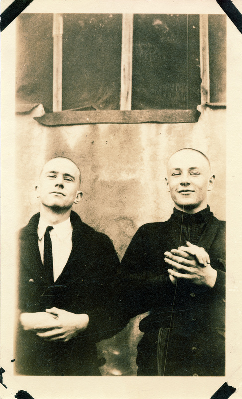 Two men pose next to each other. The back of the photograph is stamped with the number 16.