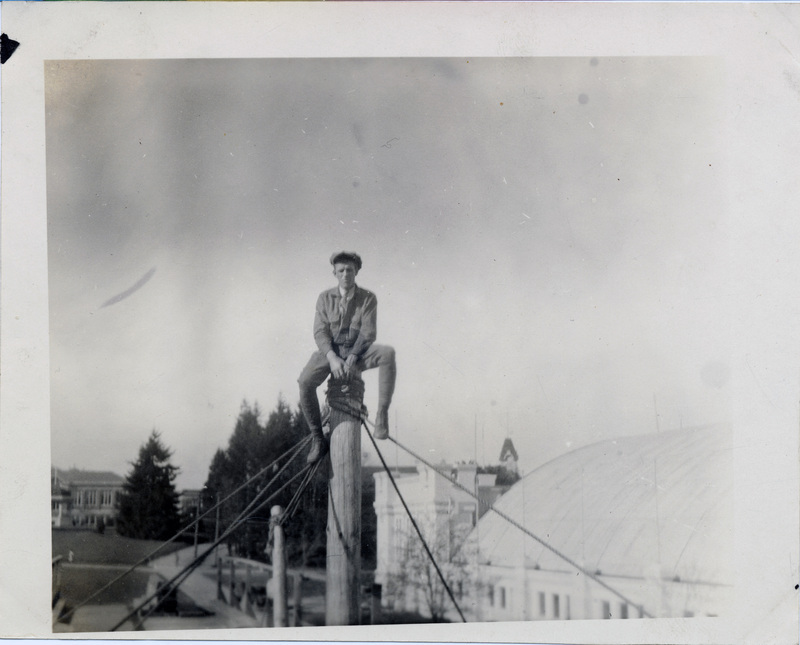 A man sits on top of a wooden pole, above the roof of a nearby building.