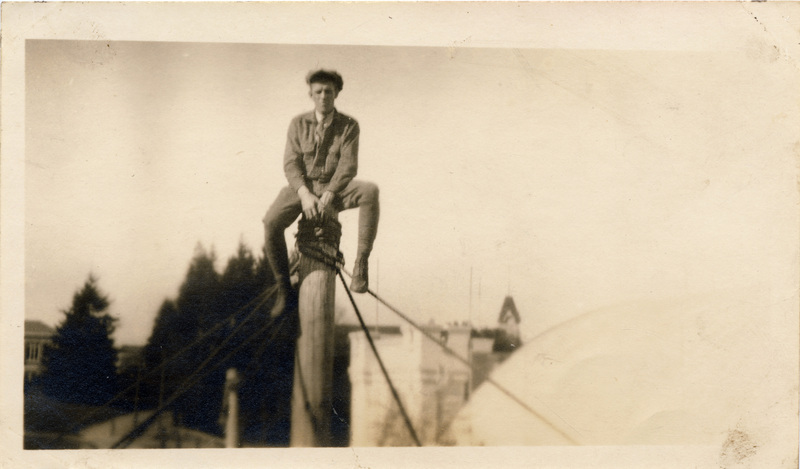 A man sits on top of a wooden pole, above the roof of a nearby building.