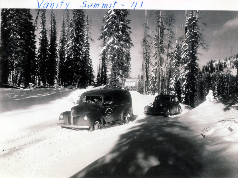 People drive two automobiles over a snowy forest road. A sign behind the vehicles reads, "Vanity Summit elevation 7700." "Vanity Summit - 1/1" is written on the front of the photograph. The back of the photograph is stamped with the number 14.