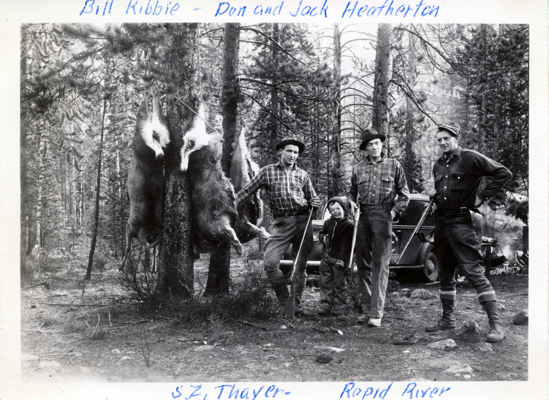 Three men and a child hold rifles and pose with deer carcasses, which are hung upside down from a tree. An automobile is visible behind them. "Bill Kibbie - Don and Jack Heatherton, S.Z. Thayer - Rapid River" is written on the front of the photograph. The back of the photograph is stamped with the number 14.