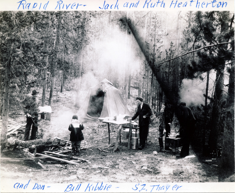 A group of people cook at a forest campsite. "Rapid River - Jack and Ruth Heatherton, and Don - Bill Kibbie - S.Z. Thayer" is written on the front of the photograph. The back of the photograph is stamped with the number 14.