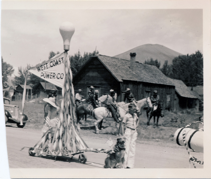 A dog pulls a parade float shaped like a lamppost with a flag reading "West Coast Power Co." A child stands next to the dog, and several people on horseback are visible in the background.