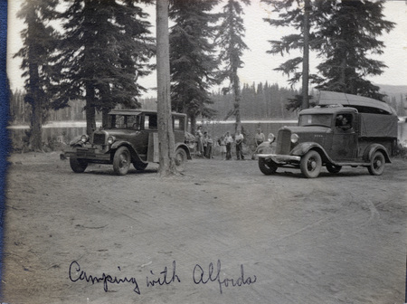 A group of people stand around two automobiles at a campsite. One automobile has a canoe tied to the top. The photo is captioned: "Camping with Alfords." On the back of the photo is written "Ready to leave Summit Lake Aug 1934."