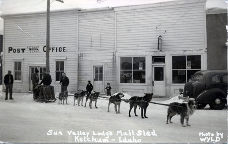 A postcard with an image of a sled dog team in front of a Post Office. On the bottom of the picture is printed: "Sun Valley Lodge Mail Sled, Ketchum - Idaho. Photo by WYLD."