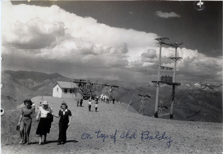 Three women wearing summer clothing walk away from a chairlift on top of a mountain. "On top of Old Baldy" is written on the front of the photograph. The back of the photograph is stamped "Jul 21 1941."