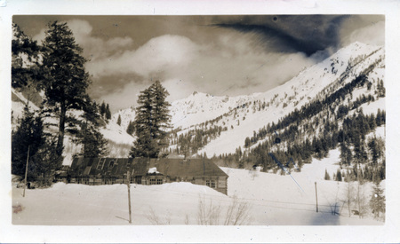 A log building at the base of a mountain, surrounded with snow. On the back of the photograph is written: "Mascot - Elev 8000 peaks back of cabin are over 10,000 Apr 17-38, Idaho."