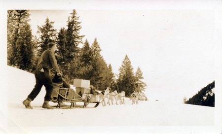 Two people and their supplies are pulled in a sled by a team of dogs. The photograph is taken from behind the sled. "Just starting after a short rest Averill took this picture. You can see what a load we had. See snowshoes taking in supplies to mine in April. Christmas presents and magazines from Nov." is written on the back of the photograph.