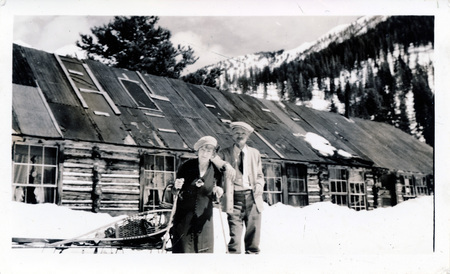A man and a woman stand next to each other in front of a single-story wooden building. Behind them is a sled with snowshoes strapped to it. "The Rutherfords Mascot Mine Idaho April 17 1938" is written on the back of the photograph.