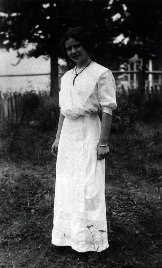 Edna wood is pictured standing in a full-length white dress with half sleeves, wearing a bead necklace and gold bracelet.