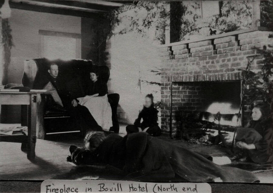 An unidentified group of people sits around a fireplace in the inside of the Bovill Hotel.