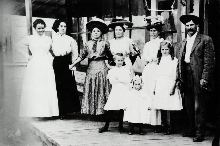 Adults, from left to right: Mrs. Alice Taylor, Mrs. Nora Harris, Mrs. George Wylie, Mrs. Jenny Price, Mrs. Case, Royal Case. In front are Opal Stockwell (right) and the smaller Case children. They are pictured standing in front of the Taylor soft drink and gaming place - actually a 'blind pig' beer parlor - which stood at Main Street and Second. Dimly seen in the background, across Pine Street, is the town recreation hall.