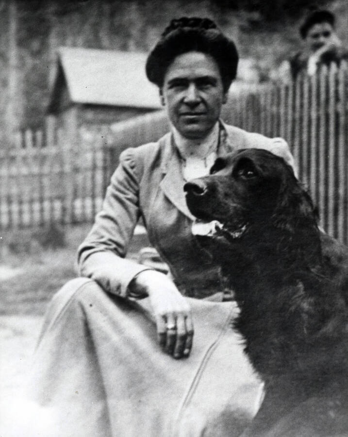 Charlotte Bovill posed seated with a dog beside a fence. In the backgroud, a person is looking over the fence toward Mrs. Bovill.