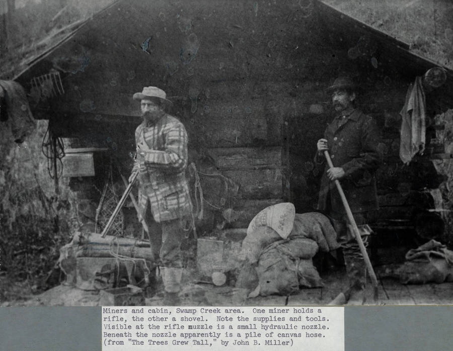 Two miners in the Swamp Creek area stand on the porch of their cabin. The man on the left is holding a rifle and standing in front of a hydraulic hose and nozzle. The man on the right is holding a shovel and is standing beside burlap sacks of supplies. From 'Trees Grew Tall.'