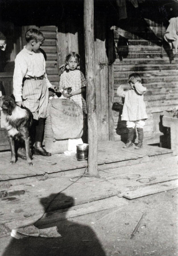 From left to right: Wallace, Naomi, and Kenneth Boll stand on a porch accompanied by a dog.
