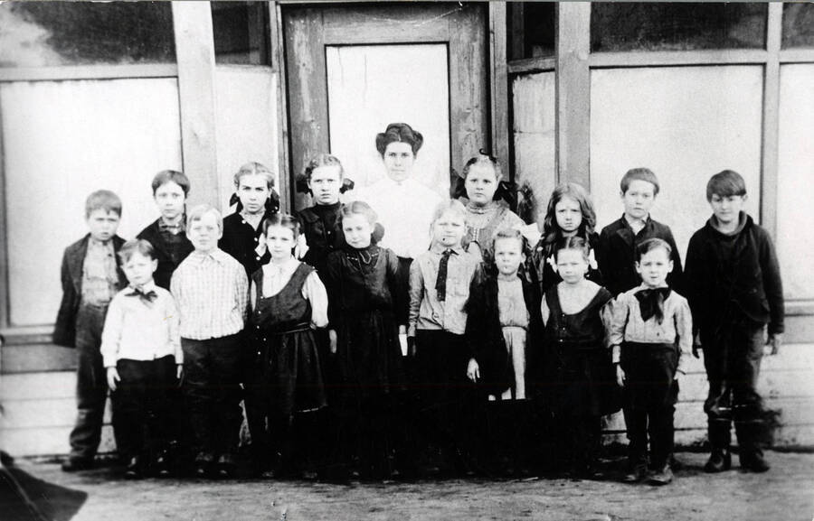 In front of Bovill's second school, located in the upper block of Main Street somewhat north of the intersection with Pine Street. This was in 1909-1910 term. Back row, starting fourth from left: Mary Smith, Miss Crane (teacher), unidentified, Opal Stockwell. Front row, starting second from left: Kneeland Parker, unidentified, unidentified, Clay Anderson (the barber's son), Helen Smith,  unidentified, Frankie Hollenbeck.