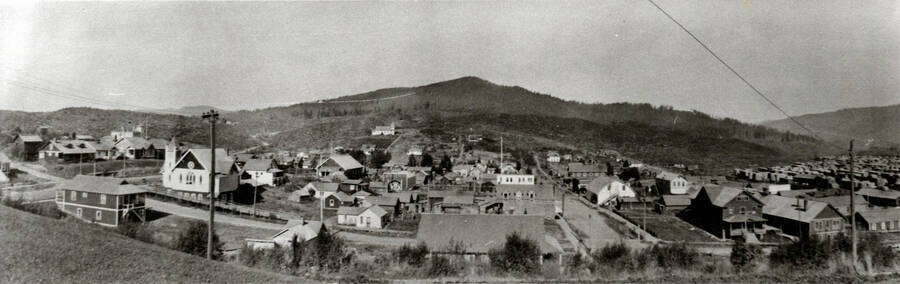 Buildings are identified in book caption: 'This is Elk River about 1915, as seen from the hill at the hospital. The prominent building toward the left, with steeple and dark gable is the Community Church. Beyond and to the left of it at the far edge of town and identified by a small belfry, is the school. The gymnasium is the large building beyond the church and to its right. Near the photo center, white and square, is the Morris Drug. Toward the camera from the drugstore is the meat market (gabled windows), which was run by Jockheck. Eastward across the street are other stores, the bank, and post office. The Catholic church stands prominently of the farther hillslope, with Shattuck Butte behind behind {sic} it. Well to the right are Potlatch buildings, the railroad tracks, and the lumber yards of the mill.'