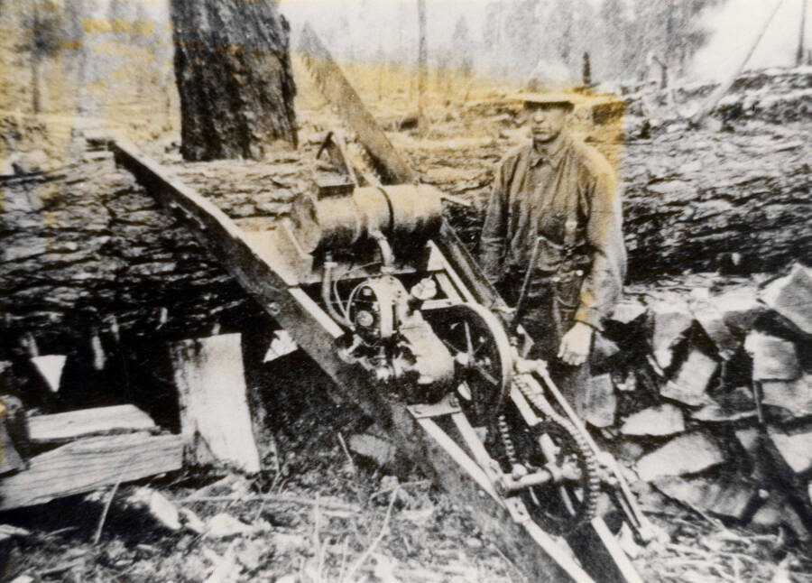 Drag-saw of the type used for wood-cutting in the 1920's. The machine was moved along the log as successive firewood blocks were cut from it. The engine was a one-cylinder, two-cycle affair. This one appears to be a Wade two-stroke dragsaw. The sawyer stands to the right of the saw.