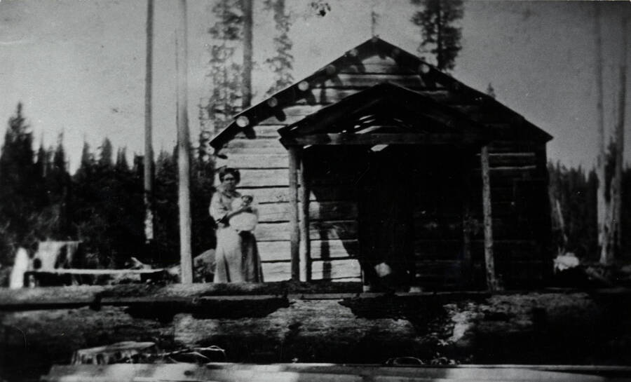 A one-room log school  house at Collins, Idaho. Mrs. Crawford, the teacher, stands next to the school.