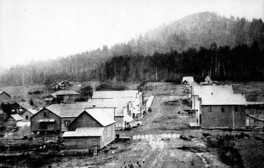 Deary about 1909, looking north across the main road, which is now the highway. The first building on the left is the Don Sahlin saloon. Beyond the corner, with a false front facing east and a gable at rear facing south, is the Bill Smith store. The buildings above are not seen clearly, but the peaked roofs of both the hardware and drug store can be distinguished, and the bank certainly existed. Farther up and to the left is the railway station, and well beyond it the Hugh Henry house. Buildings on the east (right) side of the street include the McQueary Hotel, the harness shop, and the Carlson Hotel. In the right-hand section beyond the tracks is the McGown home.