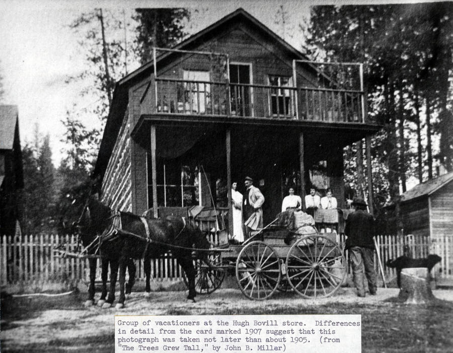 A group of visitors at the Hugh Bovill store stand on the porch. Their horse drawn carriage stands in the foreground beside a man with his back turned and a dog.