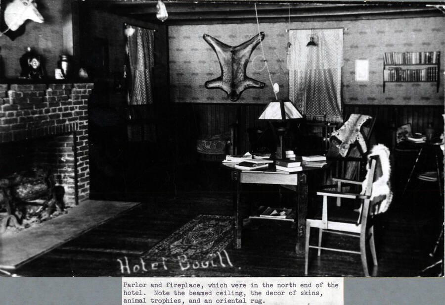 A photograph of the interior of the Bovill Hotel parlor and fireplace, locate din the north end of the building. Note the beamed ceiling and the décor of skins, animal trophies, and an oriental rug.