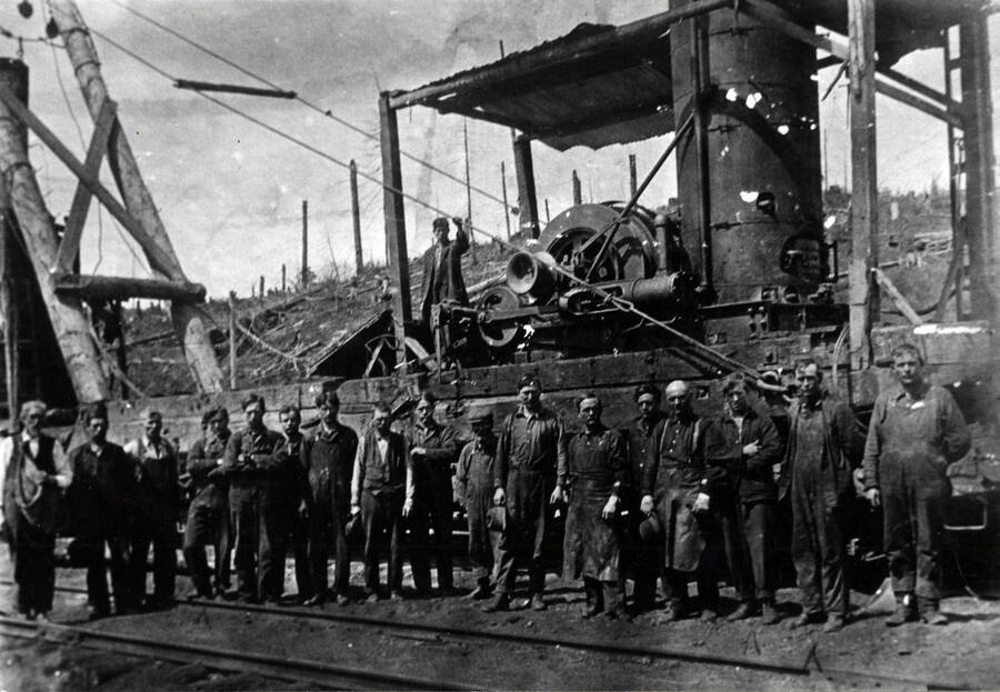 The Camp 8 crew, standing in front of a donkey engine that is rigged, evidently as a jammer type loader, on a flat-car. On the hill behind is the mess of burned timber left by the 1914 fire. In line are Grandpa Hughes, Bill Hughes, Ed Connors, Byers Sanderson; Perd Hughes, Arlie Smith, Henry Smith, Young Bill Hughes, Sammy Lowe, George Chrystal, Charlie Rist (blacksmith), Billy Coffield, Oscar Palm (blacksmith, bald head), Frank Sanderson, Andy Breeding. The man on the machine is not identified.