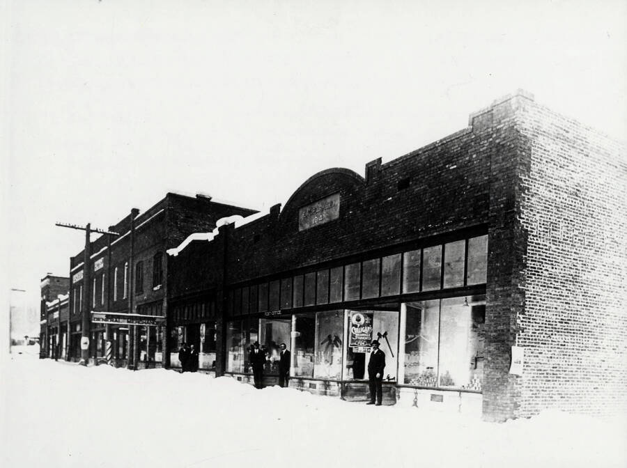 The new buildings of Main Street in January 1915. Just after reconstruction of the town. (The plan here conforms to what is shown on Map 5 in the book 'The Trees Grew Tall,' by John B. Miller). Seen in the first doorway is E.K. Parker. In the 2nd, with another man a little to one side, is Dan Featherstone. A showbill: 'Black Crow' minstrel type - The Oldest and Best Threatre - Tuesday, January 12.' Another bill on the wall of the store is readable in except for three words here reproduced as blanks: The Road to Ruin - an uplift drama with a cast of Broadway stars. The _____ _____ _____ people - 250 scenes.'