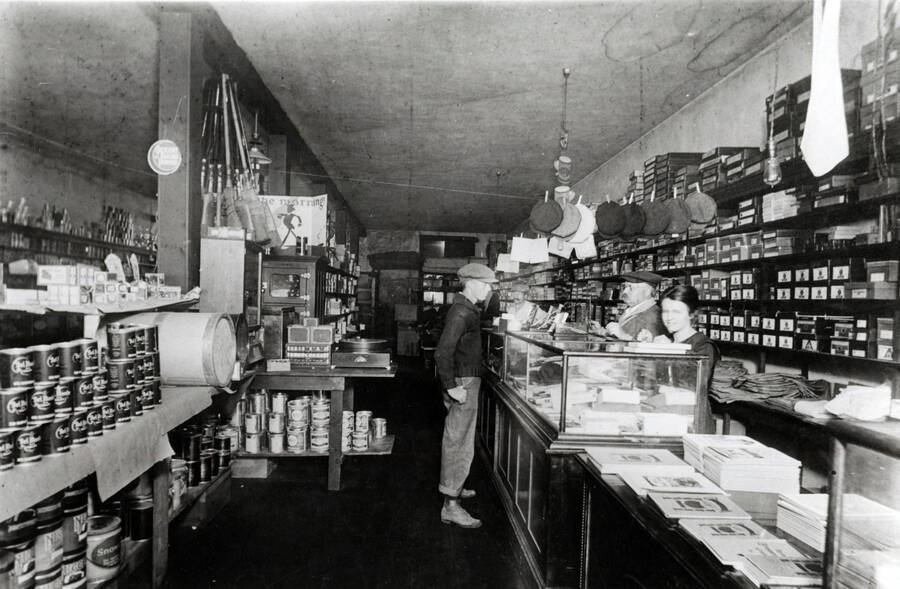 In the Groh Brother's general store. School supplies are in the forward part at the right, and dry goods behind. Groceries occupy the left hand side. Roy Perry faces the counter; Tom Groh and Marie Zagelow are behind the counter.