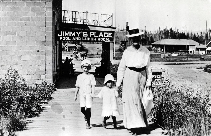 A woman and two children walk down the sidewalk in Bovill. From the caption in Trees Grew Tall: 'The view past the Verdon premises toward the depot. At this stage, the concrete building was occupied by Jimmy Gilroy.'